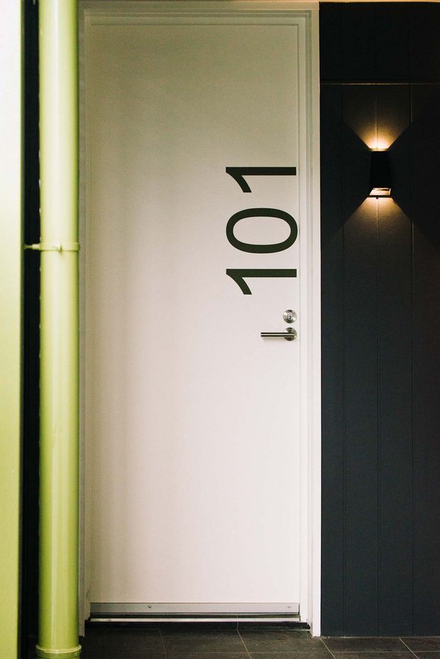 Close-up architectural photograph of a white entrance door marked with the number 107, set within a dark vertically panelled wall. A lime-green exposed pipe runs alongside the door, and a modern black-and-gold wall sconce provides directional lighting. Dark tiled flooring and clean geometric detailing create a contemporary, slightly industrial entry presentation.