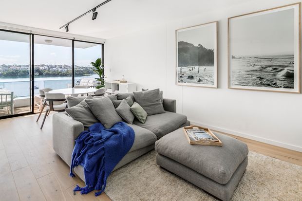 Bright open-plan living room with a large grey sectional sofa and matching ottoman, layered grey cushions, a vivid blue throw, framed coastal photography, pale timber flooring, and neutral carpeting. Floor-to-ceiling glazing opens to a balcony or terrace with a visible waterfront and urban shoreline beyond. A small dining area is positioned beside the glazing.