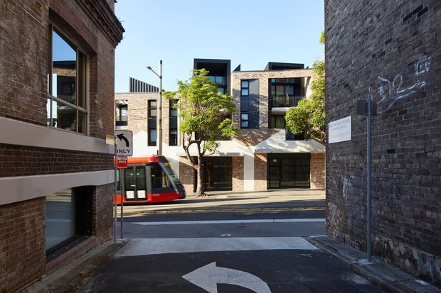 Street-level view from a narrow paved laneway toward a contemporary multi-storey brick apartment or commercial building, with a red-and-white light rail vehicle passing on the street. The surrounding buildings feature dark exposed brick façades, large black-framed windows, balconies, and projecting awnings. A historic brick building frames the left side, while the right wall carries signage and painted markings. Bright daylight and strong urban architectural context dominate the image.