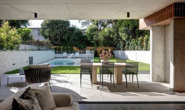 A contemporary indoor-outdoor living and dining area opening onto a landscaped backyard with a swimming pool. The covered terrace has a concrete ceiling, tiled or stone paving, a dining table with chairs, and lounge seating. Large open glazing creates a seamless connection to the lawn and pool, while exposed concrete walls and dark timber or metal detailing give the space a refined modern character.