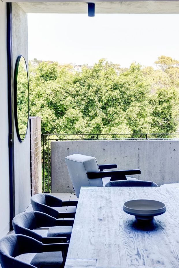 A contemporary dining area opening onto a balcony or terrace, with a long pale timber dining table, dark upholstered dining chairs, and a matching armchair. The space features a raw concrete balcony privacy wall, black metal balustrade and window/door framing, a round wall mirror, and a blue cylindrical pendant light. Dense leafy trees and neighbouring buildings are visible beyond the balcony, creating a bright indoor-outdoor setting.