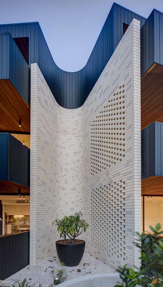 A tall, narrow architectural courtyard viewed from below, centred on a sculptural curved white brick wall with patterned breeze-block sections. Dark blue standing-seam metal cladding forms dramatic steep and concave roof or facade planes above, while warm timber soffits and recessed interior glazing flank both sides. White gravel, a curved concrete edge, and potted plants occupy the base. The image emphasises a contemporary vertical courtyard composition and strong material contrast.