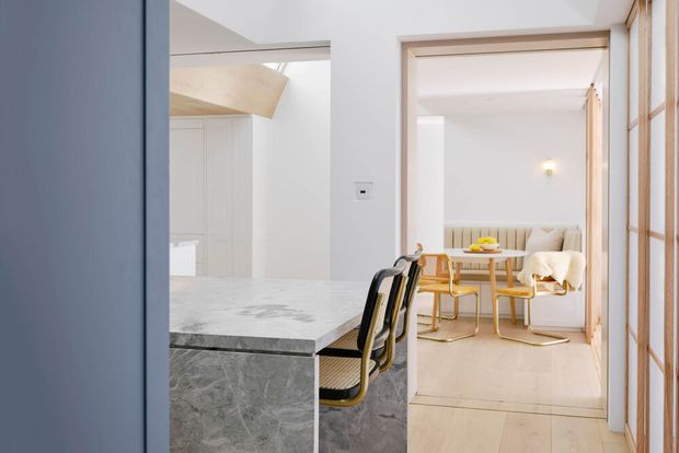 Bright contemporary interior showing a marble kitchen island with black-and-gold bar stools in the foreground, opening into a light dining area with a round table, upholstered banquette and chairs. Pale timber-framed glazing or screens and white cabinetry create an airy, refined scheme.