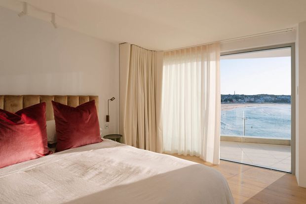 A warm, refined bedroom with a large upholstered bed, cream bedding, rich burgundy-red cushions, full-height sheer and blackout curtains, and pale timber flooring. Floor-to-ceiling glazing opens to a balcony or terrace with a clear waterfront view.
