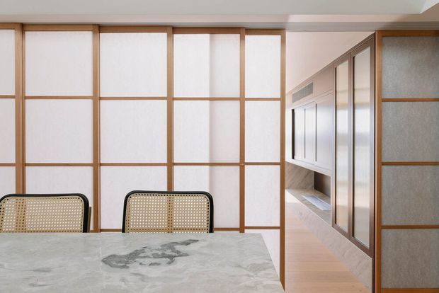A refined interior dining area featuring a pale marble dining table and woven cane dining chairs. The room is defined by extensive timber-framed translucent panel screens and sliding partitions, with matching framed glazing extending into the adjacent passage. Soft natural light, warm timber, and pale surfaces create a calm contemporary Japanese-inspired atmosphere.