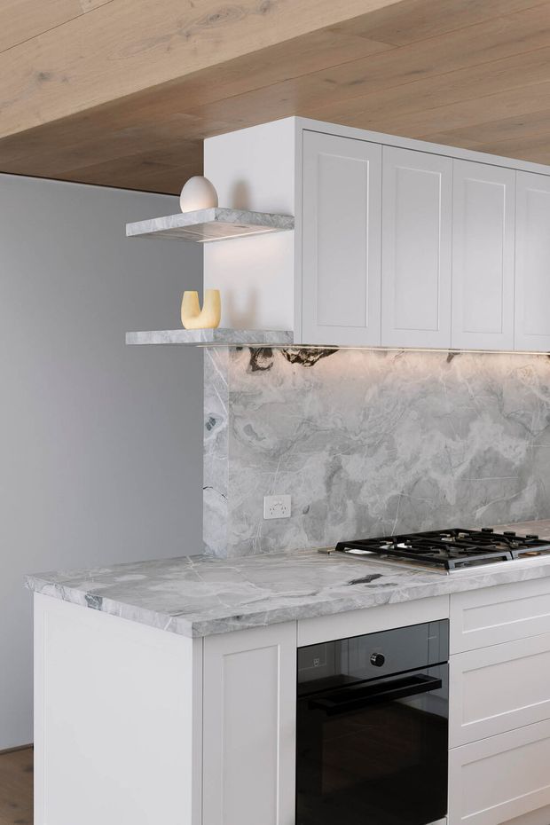 A refined kitchen featuring white Shaker-style cabinetry, a grey-veined natural stone benchtop and matching splashback, black integrated oven and gas cooktop, open stone shelves, and a warm timber-lined ceiling. The palette is light grey, white, and pale timber with subtle decorative accents.