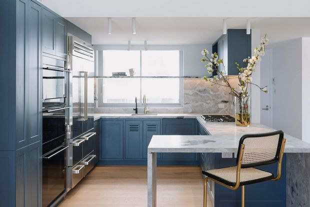 A bright, refined contemporary kitchen with deep blue-grey shaker-style cabinetry, integrated stainless-steel appliances, pale timber flooring, and a large marble-look stone benchtop extending into a dining peninsula. A matching stone splashback, expansive window, track lighting, and a cane-back chair complete the space.