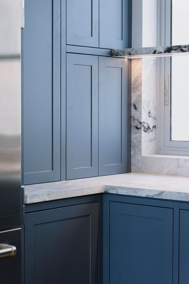 Close-up view of a compact kitchen corner with blue-grey Shaker-style cabinetry, marble-look benchtops and matching marble splashback around a window. The cabinetry includes tall upper storage and lower corner units with recessed panel doors, creating a refined, cool-toned fitted kitchen.