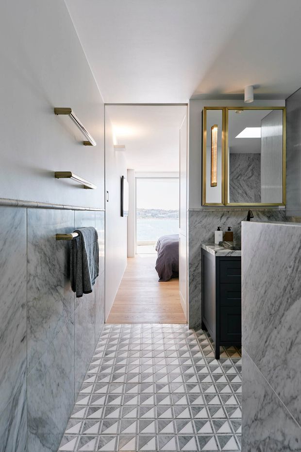 A narrow, refined bathroom with grey marble wall finishes, a geometric patterned tiled floor, brass-framed mirrored cabinetry, dark vanity joinery, and brass towel rails. An open doorway leads directly to a bedroom with a view toward water, while a skylight and wall lighting brighten the space.