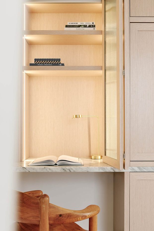 A close-up of a built-in light-wood reading nook or display cabinet with open shelving, integrated warm LED lighting, a marble ledge, books, and a small brass task lamp. An open paneled cabinet door is visible on the right, with a sculptural wooden chair positioned below.