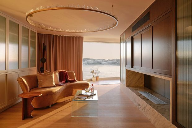 Warm, refined living room with expansive waterfront glazing, a curved caramel sofa, full-height drapery, and a large circular sculptural pendant. The room features extensive dark timber built-in cabinetry with an integrated fireplace, pale timber flooring, and a glass coffee table. The overall composition is elegant and highly resolved, with a strong indoor-outdoor connection.