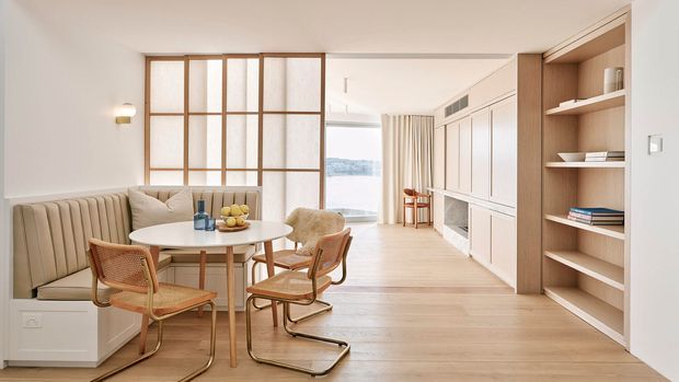 Bright open-plan dining and living area with a built-in upholstered banquette, round dining table, curved cane-and-metal dining chairs, and pale timber flooring. A timber-framed glazed partition separates the dining zone from the adjoining space, while full-height white built-in cabinetry and open shelving line the right wall. Neutral finishes, concealed storage, and soft natural light create a refined contemporary interior.