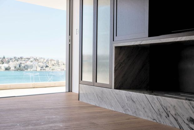A bright, modern living area with floor-to-ceiling glazing and a wide opening to a waterfront view. The room features warm timber flooring and a built-in entertainment wall finished in veined grey marble, with dark integrated cabinetry and a large television recess.