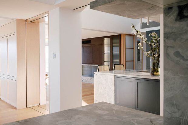 A refined contemporary open-plan kitchen and dining area viewed from behind a large stone island or benchtop. The space features pale timber flooring, white walls, dark grey built-in cabinetry, a light marble or natural-stone island with dark integrated cabinetry, timber-framed glazed joinery, and a concrete-look ceiling and foreground surface. A dining area and additional built-in storage are visible beyond the kitchen.