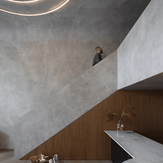 A dramatic contemporary interior featuring a sculptural staircase with a broad grey textured finish, a tall curved plaster wall, and a dark timber slatted wall beneath the stair. A marble or stone-clad counter projects into the space, while concealed circular ceiling lighting emphasizes the architectural geometry.