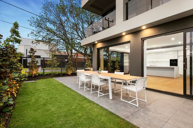A contemporary multi-level home viewed from a landscaped backyard, with a generous paved outdoor dining terrace and a long white dining table with matching chairs. Large black-framed glazed doors open to a bright white kitchen, while an upper-level balcony with glass balustrading projects above. The garden includes a manicured lawn, planted borders, mature trees, and a dark vertical-slat boundary fence. Warm recessed exterior lighting illuminates the terrace.