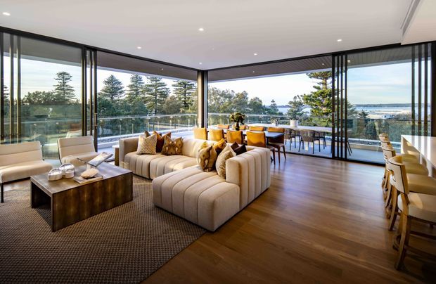 A spacious contemporary living and dining area with expansive floor-to-ceiling glazed sliding doors opening to a balcony or terrace. The room features a large cream modular sectional sofa, textured area rug, dark wood coffee table, timber flooring, and a dining table with upholstered chairs. Slim black-framed glazing and vertical black screening elements frame panoramic views of trees and water beyond. Recessed ceiling lighting and warm neutral furnishings create a refined, inviting interior.