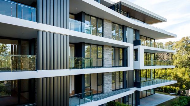 Exterior view of a contemporary multi-storey residential building with strong horizontal slab projections, dark vertical ribbed cladding, extensive floor-to-ceiling glazing, glass-balustraded balconies, and sections of light stone wall cladding. The building is set among mature trees and landscaped greenery.