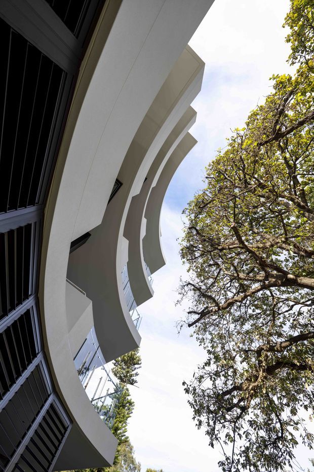 Upward-looking exterior architectural photograph of a multi-storey building with strong curved white projecting balconies and dark vertical window or louvre elements. Mature trees frame the right side, while the upward perspective emphasises the building's sculptural facade and layered cantilevered forms.