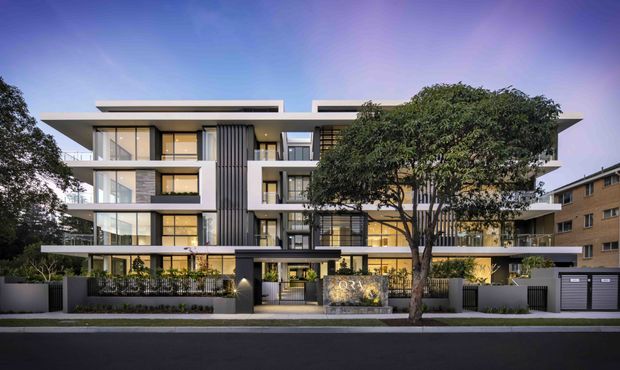 Street-facing view of a contemporary multi-storey apartment building with a layered flat roofline, projecting balconies, extensive glazing, dark vertical screening, and light rendered facade elements. Mature street trees and landscaped planting frame the front entry, with a neighbouring apartment building visible to the side.
