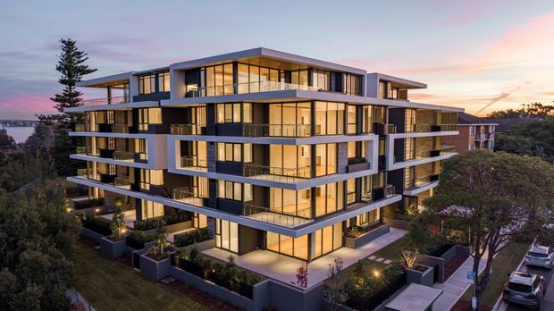Exterior architectural photograph of a large contemporary multi-storey apartment building at dusk. The building features stacked projecting balconies, extensive floor-to-ceiling glazing, dark and light facade panels, flat rooflines, landscaped communal edges, and warm interior lighting. Residential surroundings and parked cars are visible in the background.