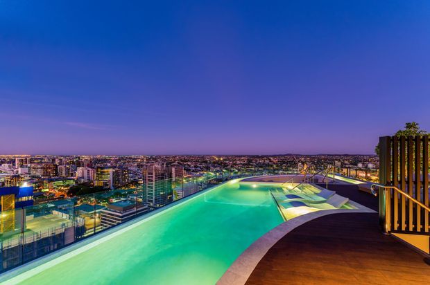 A twilight rooftop terrace featuring a large illuminated swimming pool, timber decking, glass balustrades, and a city skyline stretching across the horizon. The scene has a luxurious resort-like atmosphere with dramatic blue and purple evening light and integrated outdoor lighting.