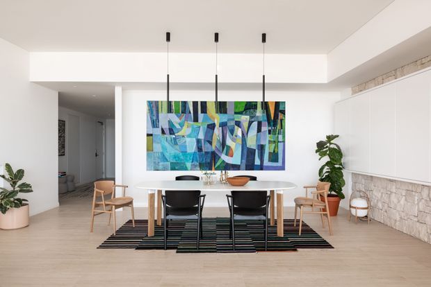 Bright contemporary dining area with a white oval dining table, mixed black and timber dining chairs, three black pendant lights, and a large colourful abstract artwork. The room features pale timber flooring, white walls and ceiling, extensive white built-in cabinetry, and a textured light stone feature wall. Indoor plants and restrained furnishings create a clean, modern atmosphere.