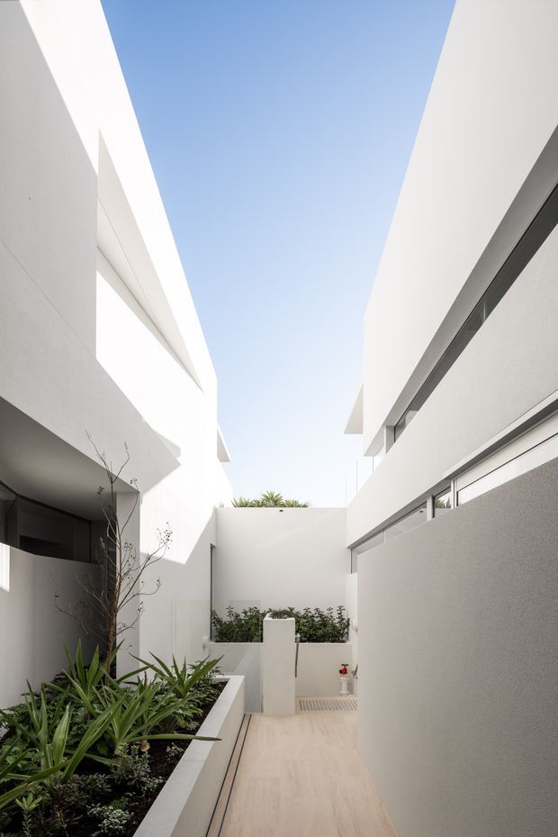 A narrow, minimalist architectural courtyard or external walkway between crisp white rendered building volumes. The passage has pale tiled paving, integrated raised planters with drought-tolerant greenery, concealed or linear glazing, and strong geometric overhangs framing an open strip of blue sky. The scene is bright, clean, and contemporary with high vertical walls and carefully controlled natural light.