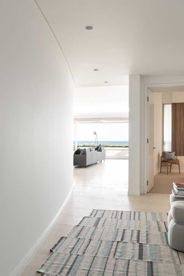 Bright minimalist residential hallway opening into a light-filled living room with expansive ocean views. The corridor has smooth white walls, recessed ceiling lights, pale timber flooring and a large muted geometric runner. An open doorway reveals a softly furnished bedroom or sitting room with a timber panel feature and upholstered chair. The distant living area contains a grey sofa and floor lamp, with broad glazing facing the sea.