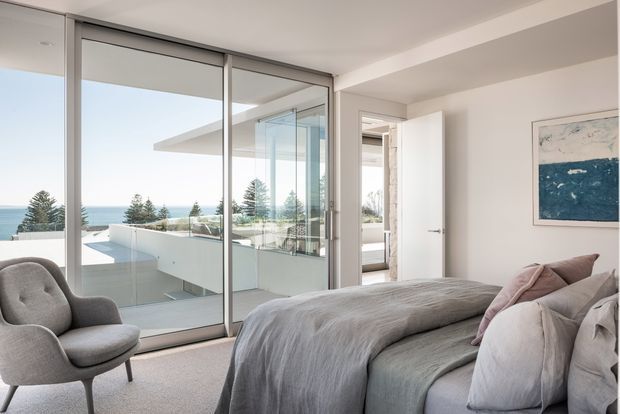 Bright contemporary bedroom with a large upholstered bed, grey bedding, plush neutral armchair, and full-height sliding glass doors opening to a balcony or terrace. The glazing provides expansive coastal ocean views, while white walls and grey finishes create a calm, refined interior. A partially open door leads to an adjacent space, and abstract blue artwork adds a subtle accent.
