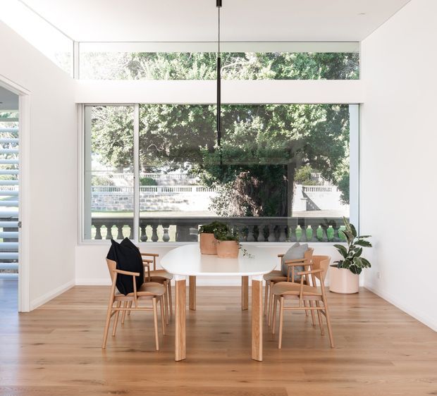 Bright, minimalist dining area with a round white dining table and four timber dining chairs, set against large floor-to-ceiling windows. The room has white walls, a white ceiling, warm timber flooring, simple black pendant lighting, and potted plants. Exterior greenery and a balustrade are visible through the glazing.