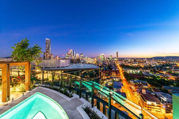 Twilight rooftop terrace overlooking a brightly lit urban skyline, featuring a turquoise swimming pool, glass balustrades, landscaped planting, and a contemporary glass-and-metal rooftop structure. The elevated city outlook and outdoor architectural setting dominate the image.