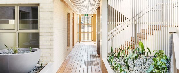 A bright contemporary residential passageway or entry corridor with pale brick walls, light timber decking, a slatted timber ceiling, and a white vertical batten screen enclosing a staircase. Built-in planter beds with white gravel and greenery line the corridor, while glazed doors and a timber security gate appear at the far end.