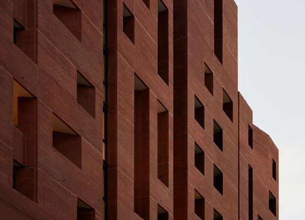 Close-up exterior view of a large contemporary brick-clad building, featuring repetitive geometric recessed window openings and deep vertical facade reveals. The dark red-brown masonry has a finely textured horizontal coursing, creating a monolithic and strongly patterned architectural envelope against a pale sky.