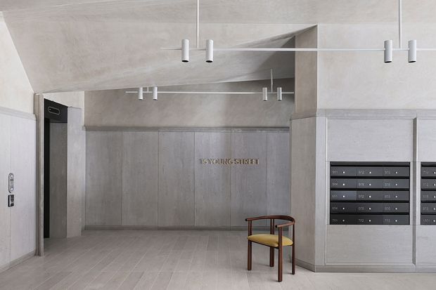 A refined apartment or building entry lobby with a minimalist concrete-toned interior. The space features pale grey architectural surfaces, integrated wall panels, a tiled floor, recessed and linear ceiling geometry, slim pendant lighting, apartment mailboxes, an elevator entrance, and a single timber-framed chair with a mustard upholstered seat. A small metallic wall sign is centered on the rear panel.