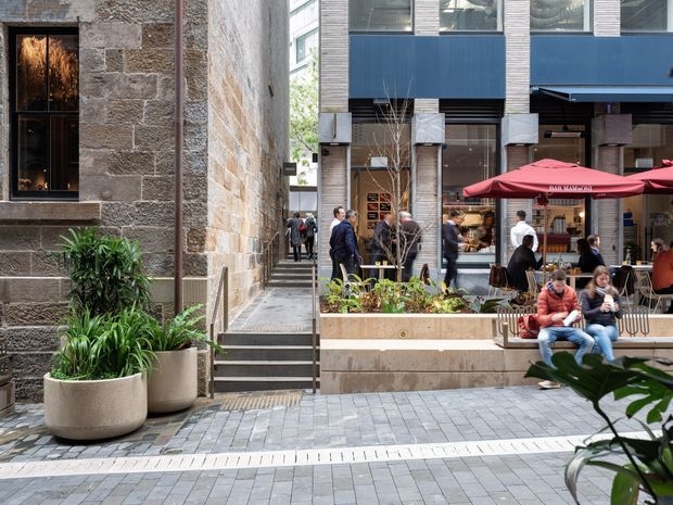 Urban commercial laneway or courtyard viewed at pedestrian level, with a historic rough stone masonry building on the left beside a contemporary multi-storey commercial facade. The space includes broad grey tiled paving, steps, raised planter beds with greenery, café seating, people, and a red outdoor umbrella. The architecture combines heritage masonry with modern blue-clad and glazed construction.