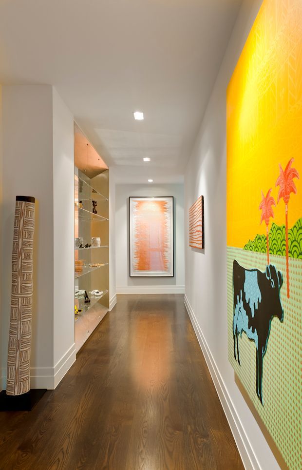 A long, refined residential hallway or gallery corridor with dark timber flooring, white walls and ceiling, recessed downlights, and built-in glass display shelving. Framed artwork and a large vibrant patterned artwork create colourful focal points along the passage, with a sculptural freestanding piece near the entrance.