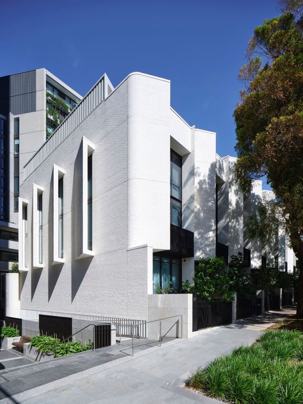 Street-level exterior view of a contemporary multi-storey residential building with a strong white brick facade, tall deeply recessed vertical windows, projecting geometric volumes, dark metal balcony and fence elements, and a stepped roofline. The building fronts a paved pedestrian walkway with landscaped planting and mature trees under a clear blue sky.