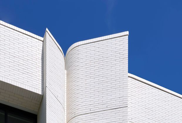 Close-up exterior architectural view of a contemporary building featuring crisp white brick cladding, intersecting curved and angular wall forms, and strong geometric rooflines against a deep blue sky. The image emphasizes the sculptural facade and masonry detailing rather than doors, windows, or interior spaces.