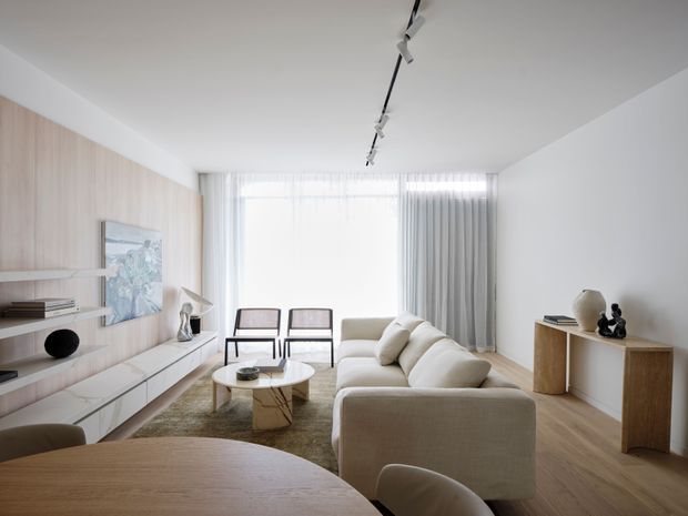 Bright, minimalist contemporary living room viewed from the dining area. The room features pale timber wall panelling and flooring, a long built-in media/storage unit, a large neutral upholstered sofa, a round coffee table, restrained decor, sheer curtains with grey drapes, and a linear black track light. A dining table and chairs occupy the foreground.