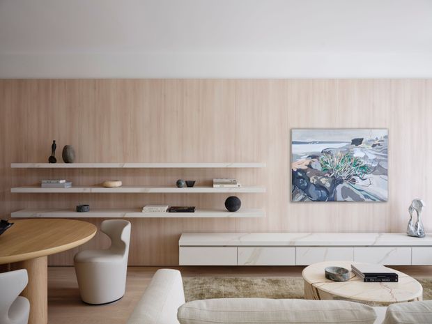Contemporary living room with a warm light-wood feature wall, floating open display shelves, and a long white built-in media cabinet with a stone-look surface. A curved dining table and upholstered chairs sit to the left, while a pale sectional sofa and round coffee table occupy the foreground. The space is softly lit and minimally styled with neutral furnishings, sculptural objects, and abstract artwork.