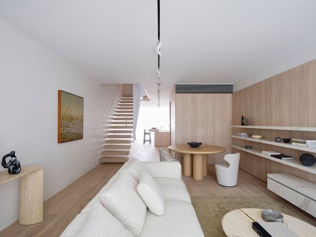 Bright, refined open-plan living room with a white sectional sofa, round timber dining table, and extensive light timber built-in shelving and cabinetry. A sculptural open-tread staircase leads toward the rear of the home, while a linear black track light runs along the white ceiling. The palette is predominantly white and light wood, with a calm contemporary feel.