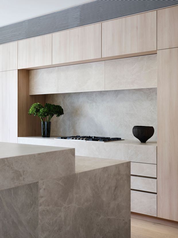 A refined contemporary kitchen with a concealed cooking wall and large sculptural island. Pale timber cabinetry frames light stone-look panels and benchtops, while a dark gas cooktop, black decorative vessel, and greenery provide restrained contrast. A linear ceiling ventilation grille runs across the top of the cabinetry, and warm timber flooring complements the calm neutral palette.