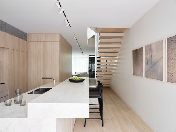 Bright, open-plan contemporary kitchen and dining area with a long white stone island, integrated light timber cabinetry, black bar stools, pale timber flooring, track lighting, and a floating timber staircase leading to an upper level. Minimal abstract wall art and a softly screened living area are visible in the background.