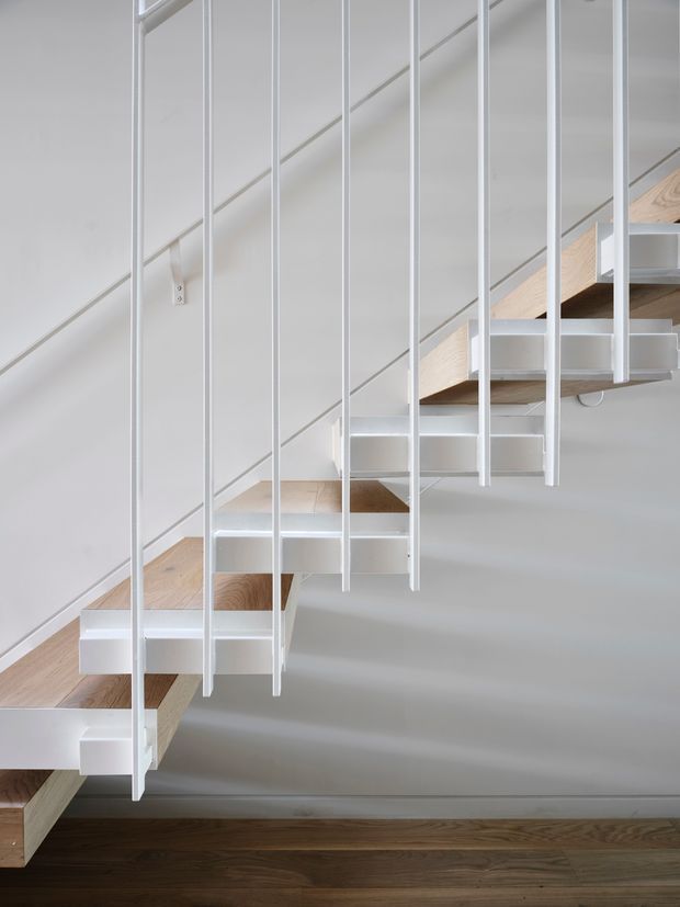 Close-up interior view of a minimalist floating staircase beneath a sloped ceiling. The stair treads are light timber with white structural supports and numerous slender vertical white metal balusters, set against white walls and a dark timber floor.