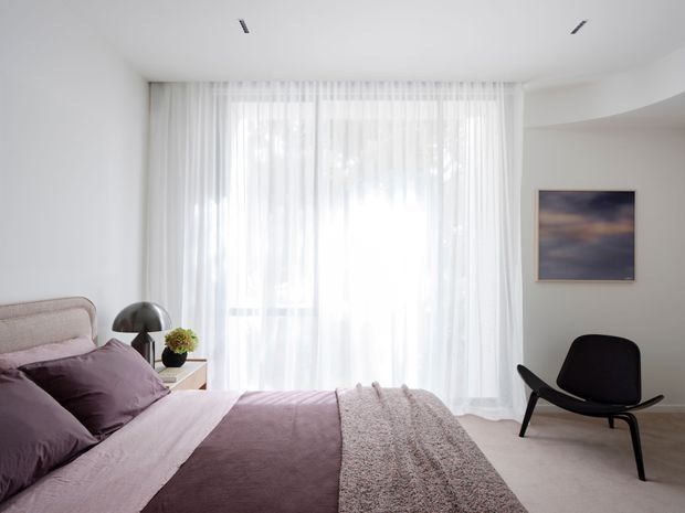 Bright, softly styled contemporary bedroom with a large floor-to-ceiling window dressed in sheer white curtains, a low upholstered bed in muted mauve and lavender bedding, a black sculptural lounge chair, bedside table and lamp, and a restrained abstract artwork. The room has white walls, recessed ceiling lighting, and a calm neutral palette with gentle natural light.