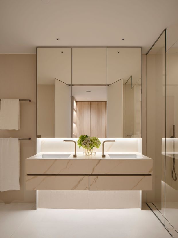 A refined, contemporary bathroom featuring a large double-vanity with integrated basins, pale veined stone surfaces, illuminated mirror cabinetry, warm beige wall finishes, and a frameless glass shower enclosure. Soft concealed lighting and neutral materials create a calm, luxurious atmosphere.