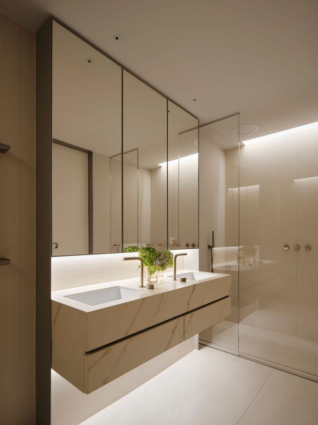 A refined contemporary bathroom featuring a floating double vanity with a pale veined stone finish, integrated basins, brushed metal tapware, full-height mirrored cabinetry, and a frameless glass walk-in shower. Warm concealed LED lighting, soft beige finishes, and vertical wall detailing create a calm, luxurious atmosphere.