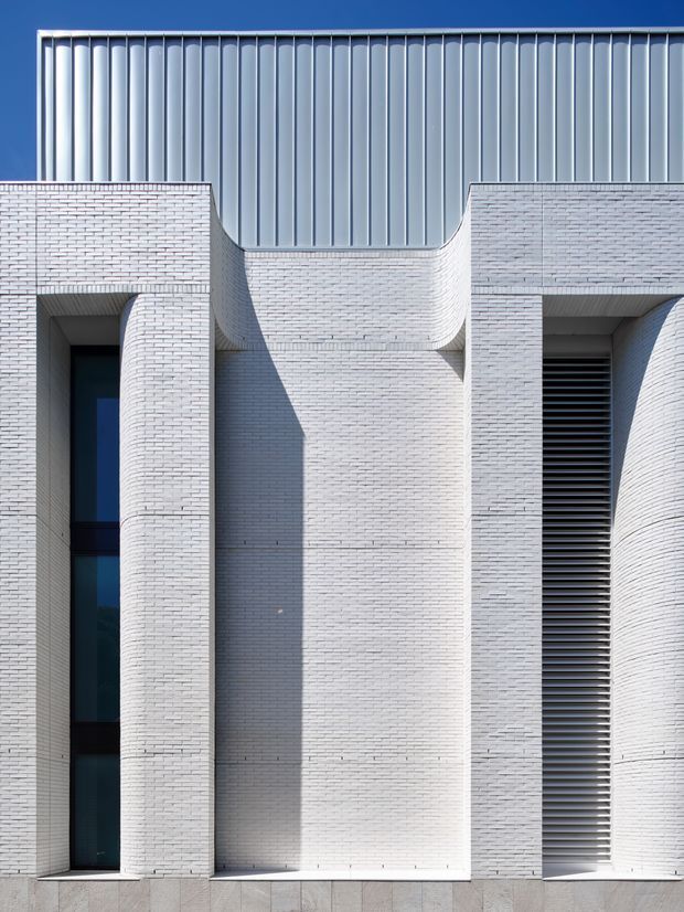 Close-up exterior view of a contemporary building facade composed of pale white brickwork, deep recessed openings, curved brick forms, dark glazing, and a prominent upper section clad in vertically ribbed silver-grey metal panels. Strong sunlight creates geometric shadows across the facade.