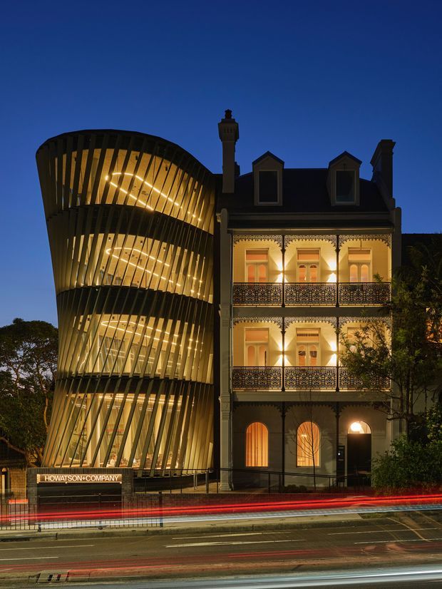 Twilight exterior photograph of a heritage-style multi-storey building beside a dramatic contemporary curved addition. The original building has a dark pitched roof, dormer windows, ornate balconies, decorative trim and illuminated arched ground-floor openings. The adjacent addition features sweeping, vertically ribbed dark framing with warm interior lighting across several levels. The street in front shows long-exposure vehicle light trails.
